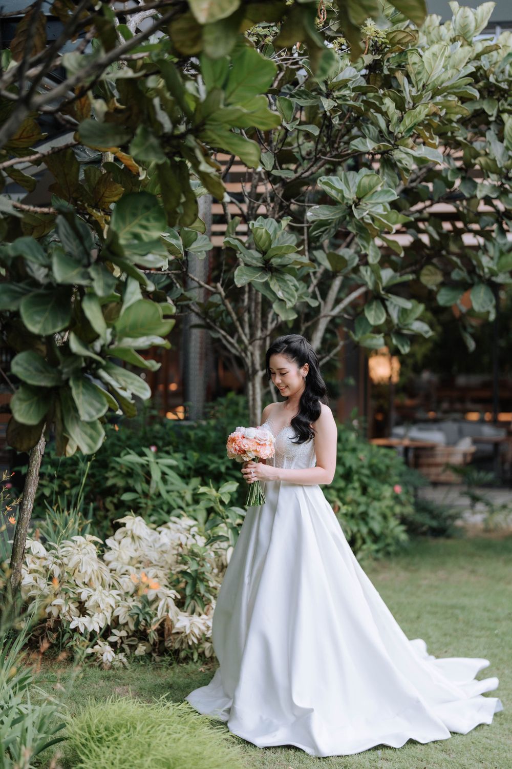 Elegant bride in white gown holds bouquet while standing in lush garden setting.