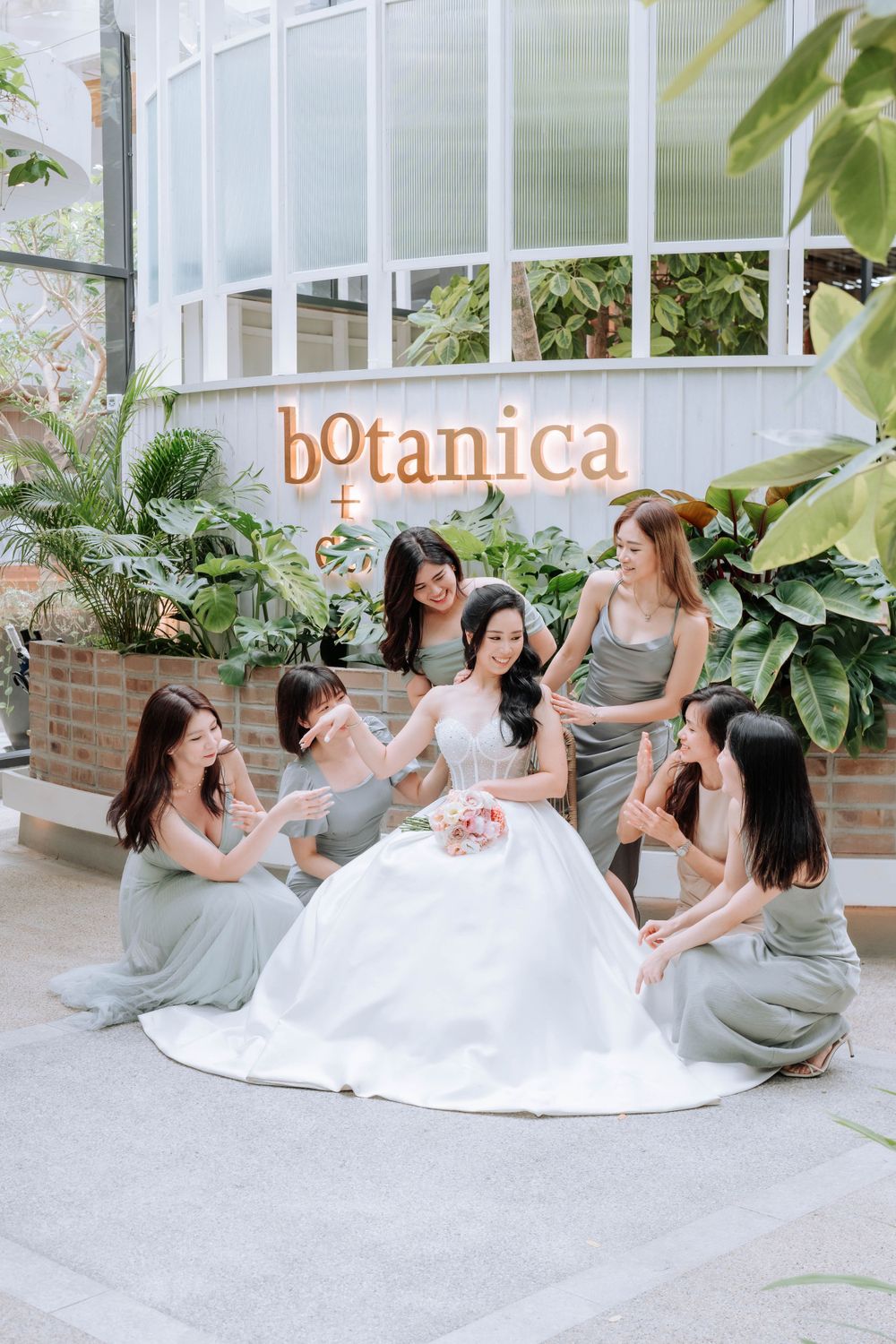 Bridesmaids in sage green dresses gather around bride at Botanica venue.