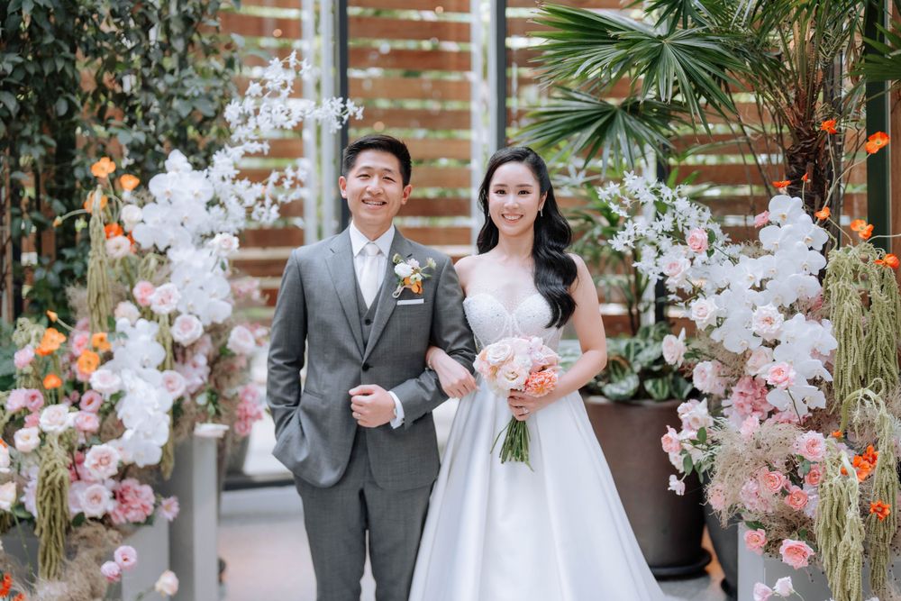 Couple stands before elegant white floral arch display at wedding venue.