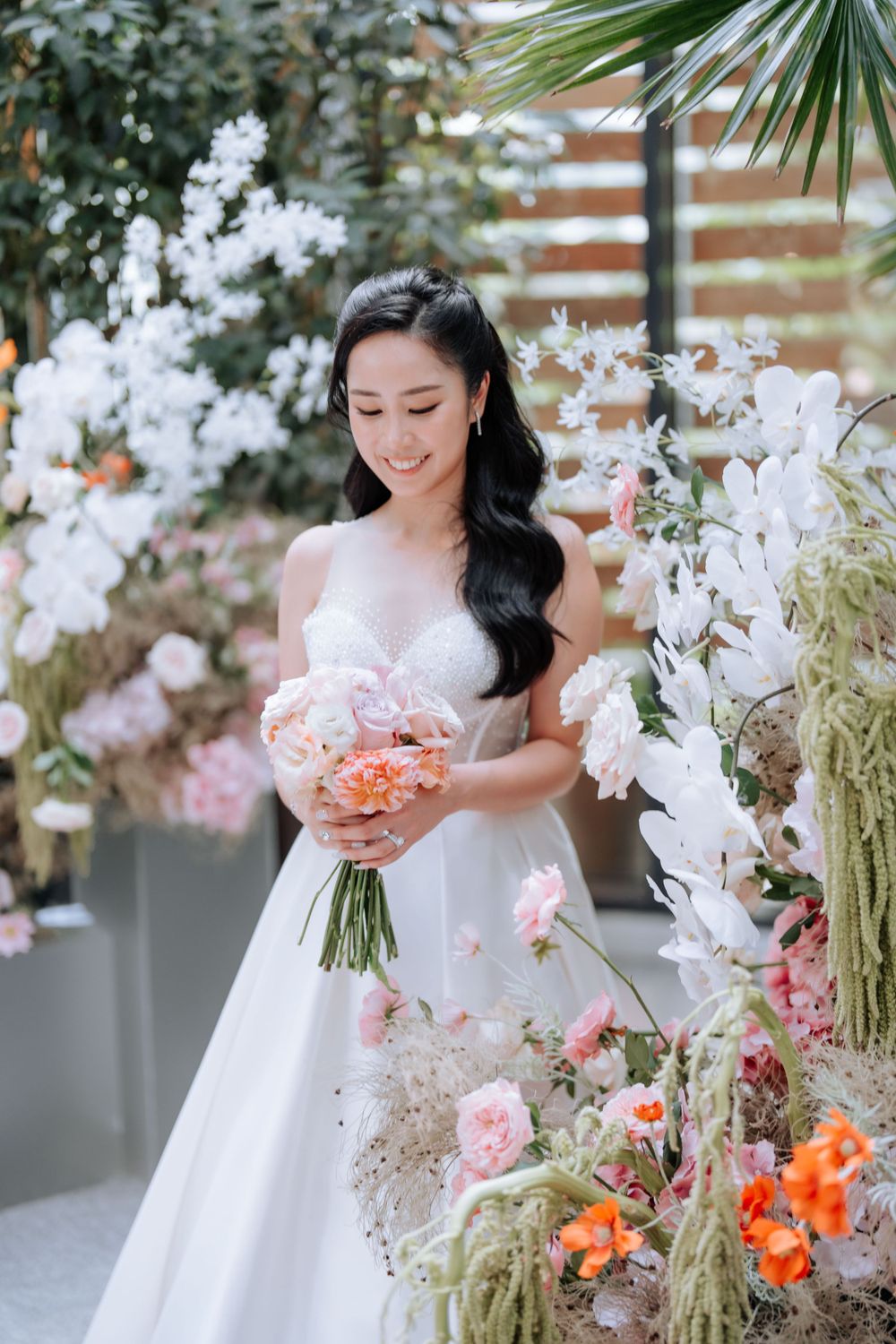 Bride with elegant white gown holds pink bouquet amid floral decorations.
