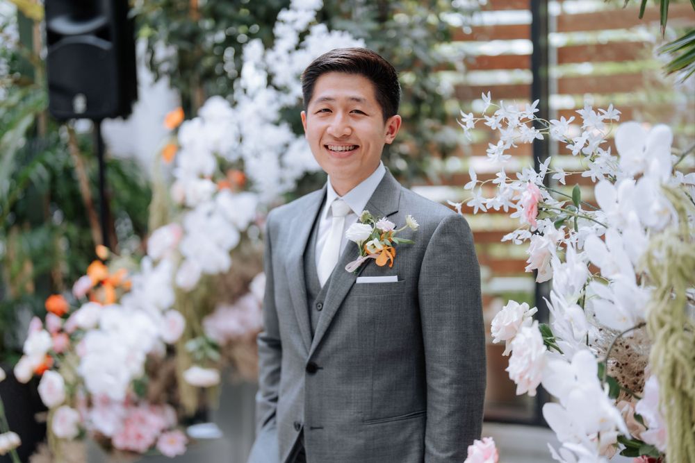 A groom in a grey suit stands smiling in front of a wedding arch decorated with white flowers and greenery.