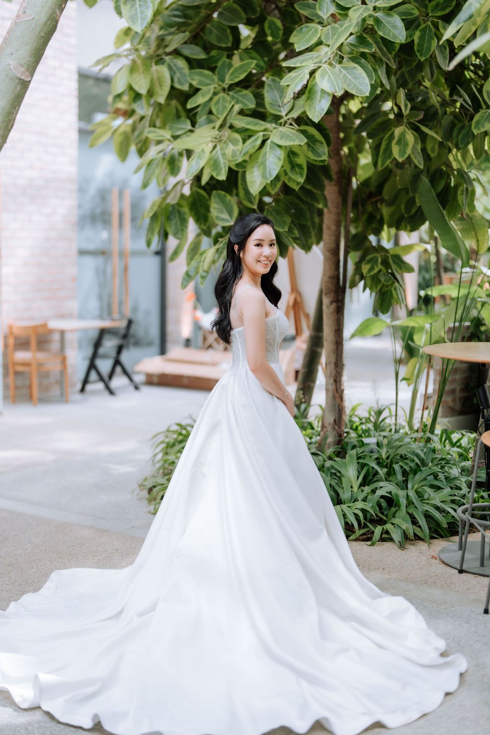 A bride in a white ballgown wedding dress poses outdoors under a lush green tree.