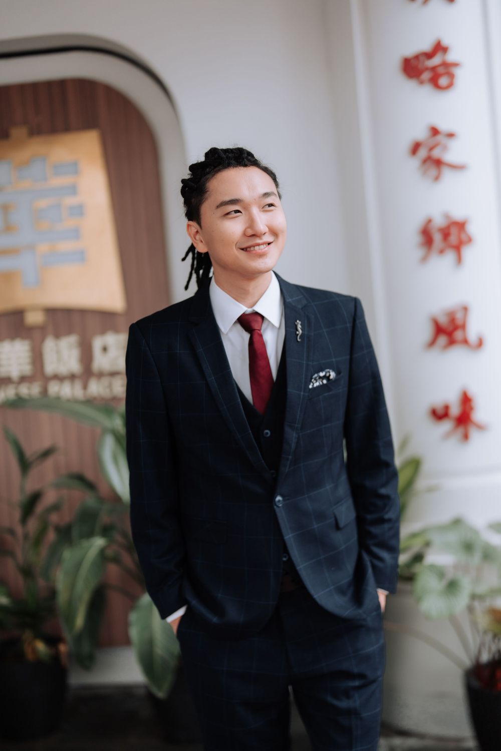 A formal portrait of a person in a navy suit and red tie standing in front of Chinese calligraphy.