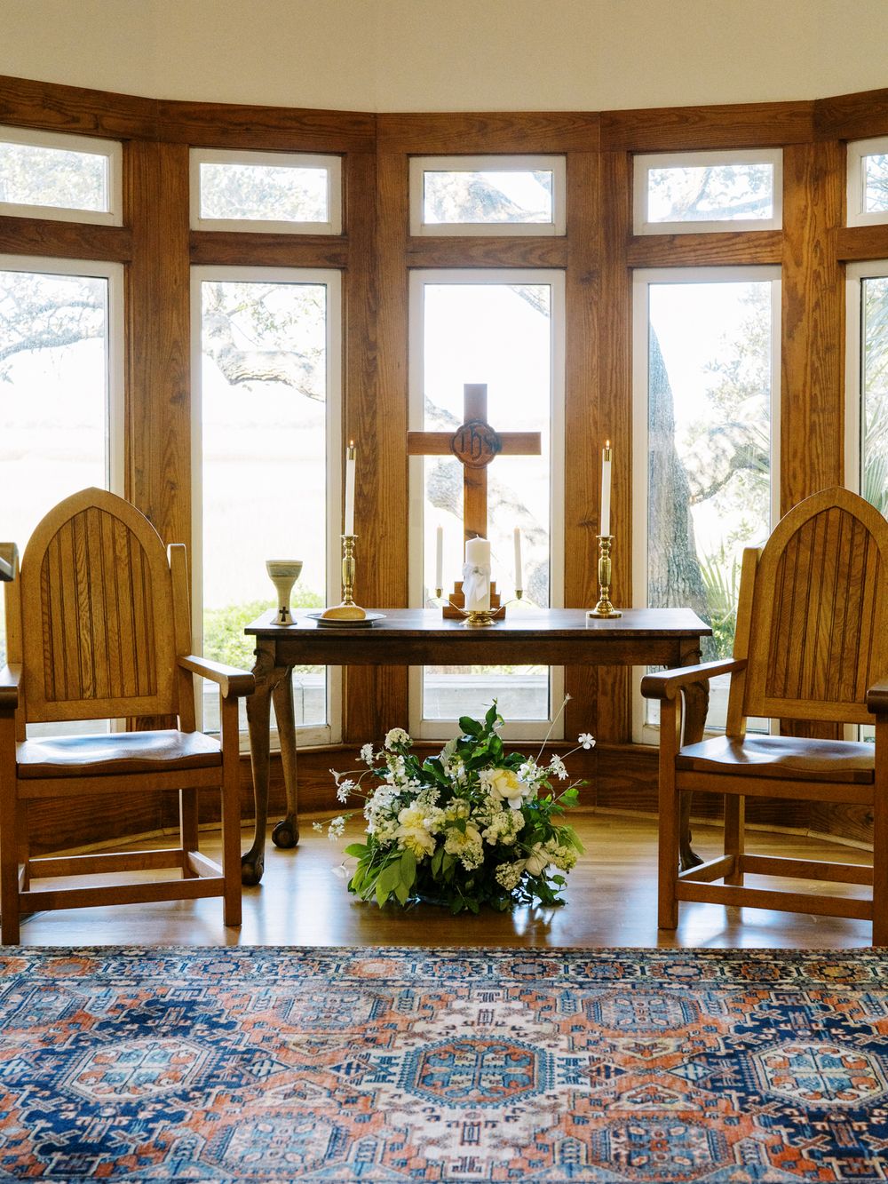 Church altar area with wooden chairs and cross surrounded by windows and floral arrangements.