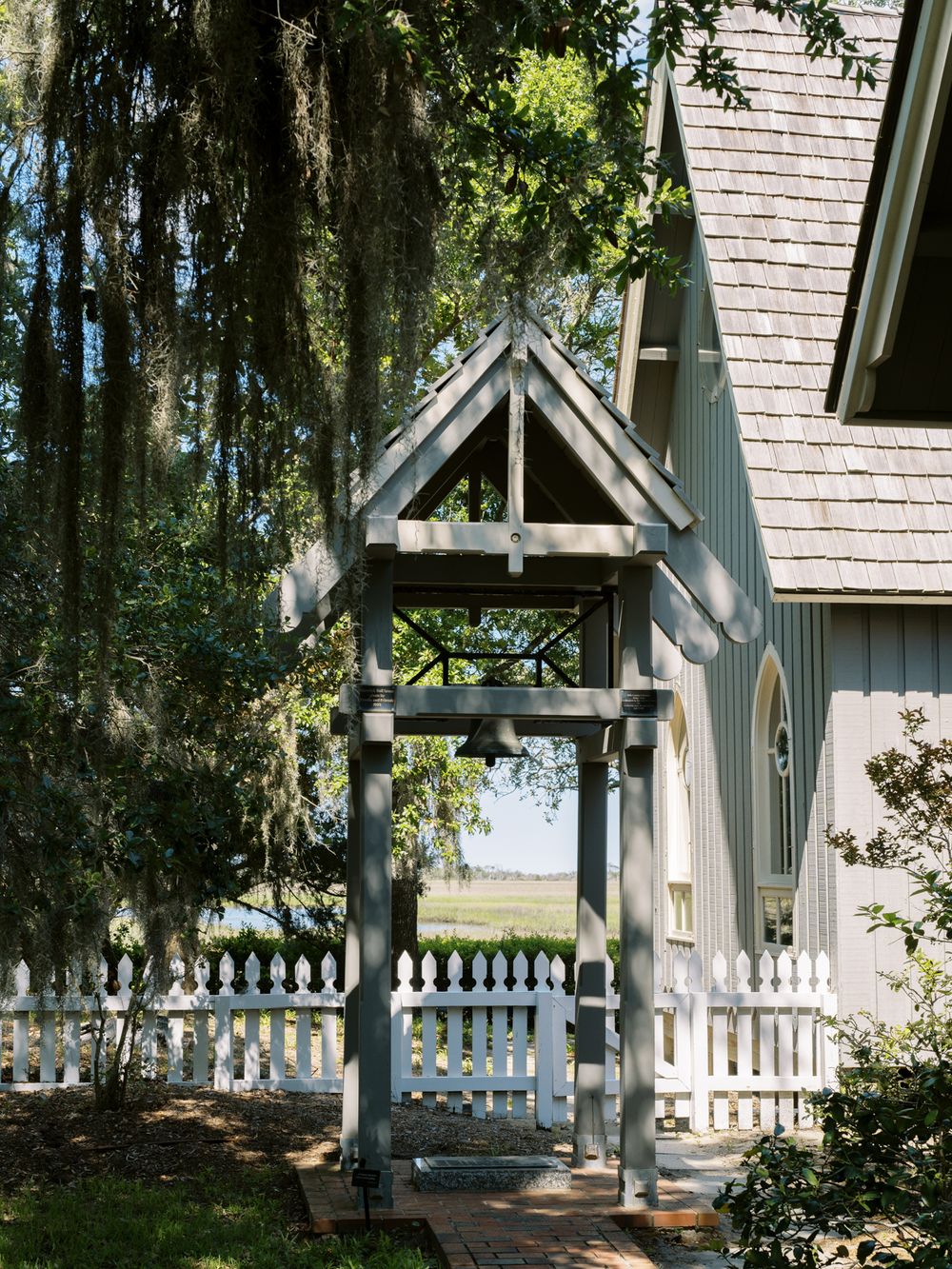 A charming white chapel entrance with a wooden archway and picket fence surrounded by trees.
