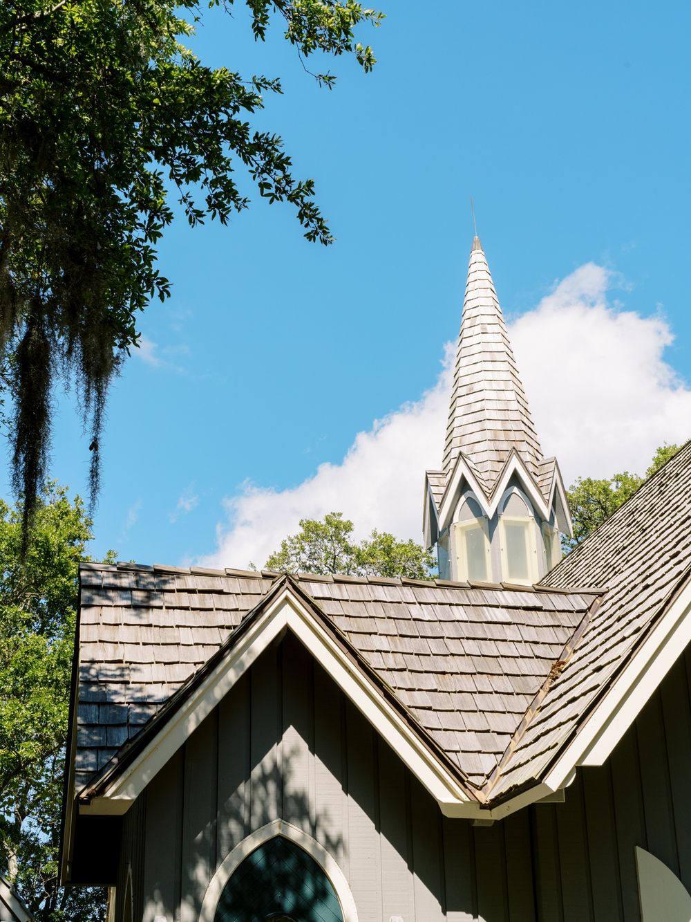 A white church spire reaches into blue sky with Spanish moss hanging from nearby trees.