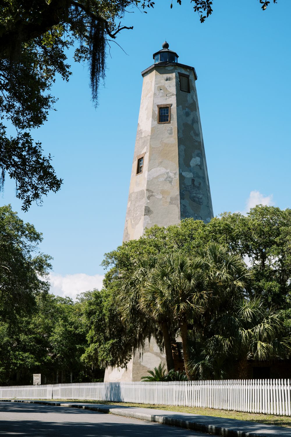 Tall white lighthouse tower surrounded by palm trees and Spanish moss in bright sunlight.