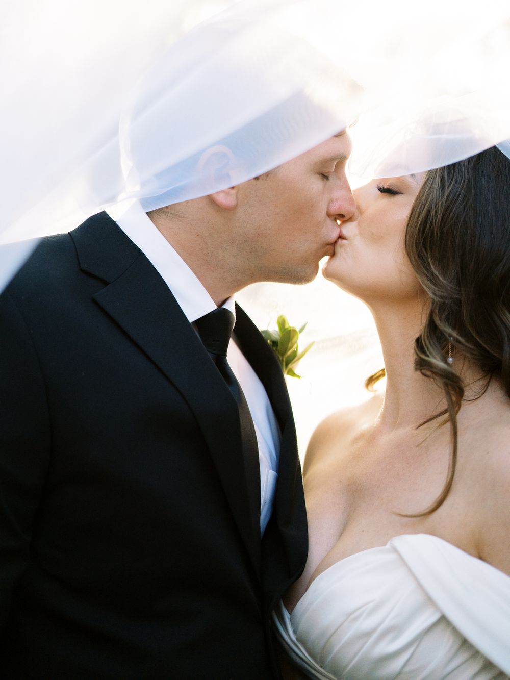 Romantic wedding kiss between couple in formal attire with sunlit background.