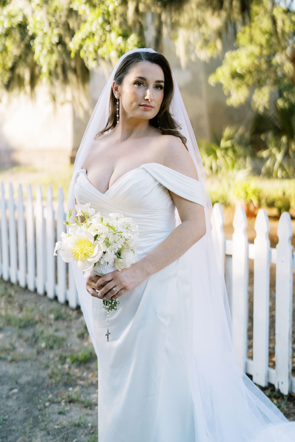 Bride in off-shoulder wedding dress holds white bouquet by white picket fence.