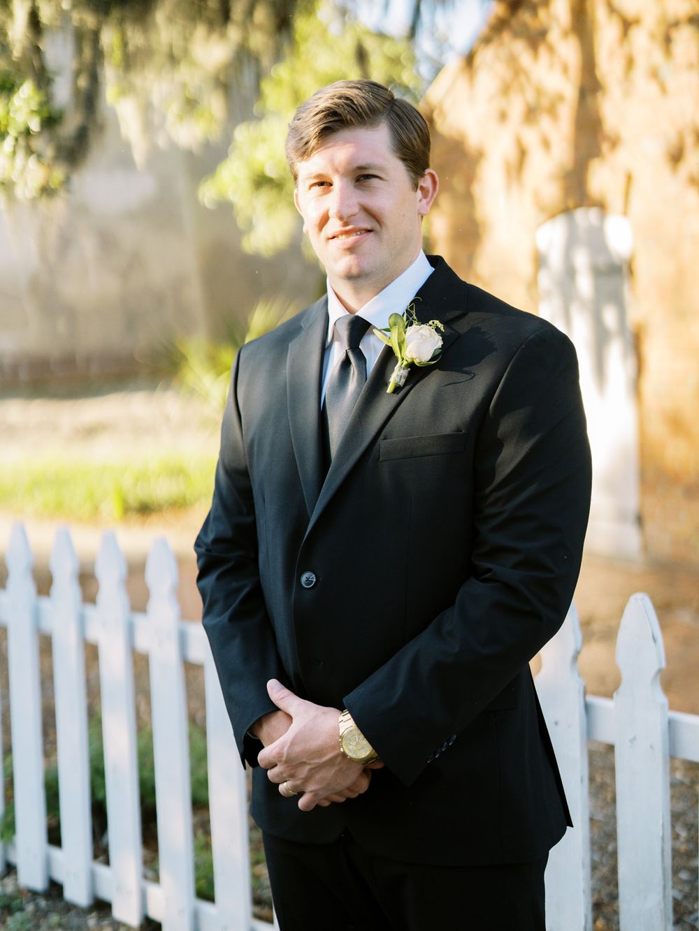 Groom in black tuxedo with boutonniere stands by white picket fence.