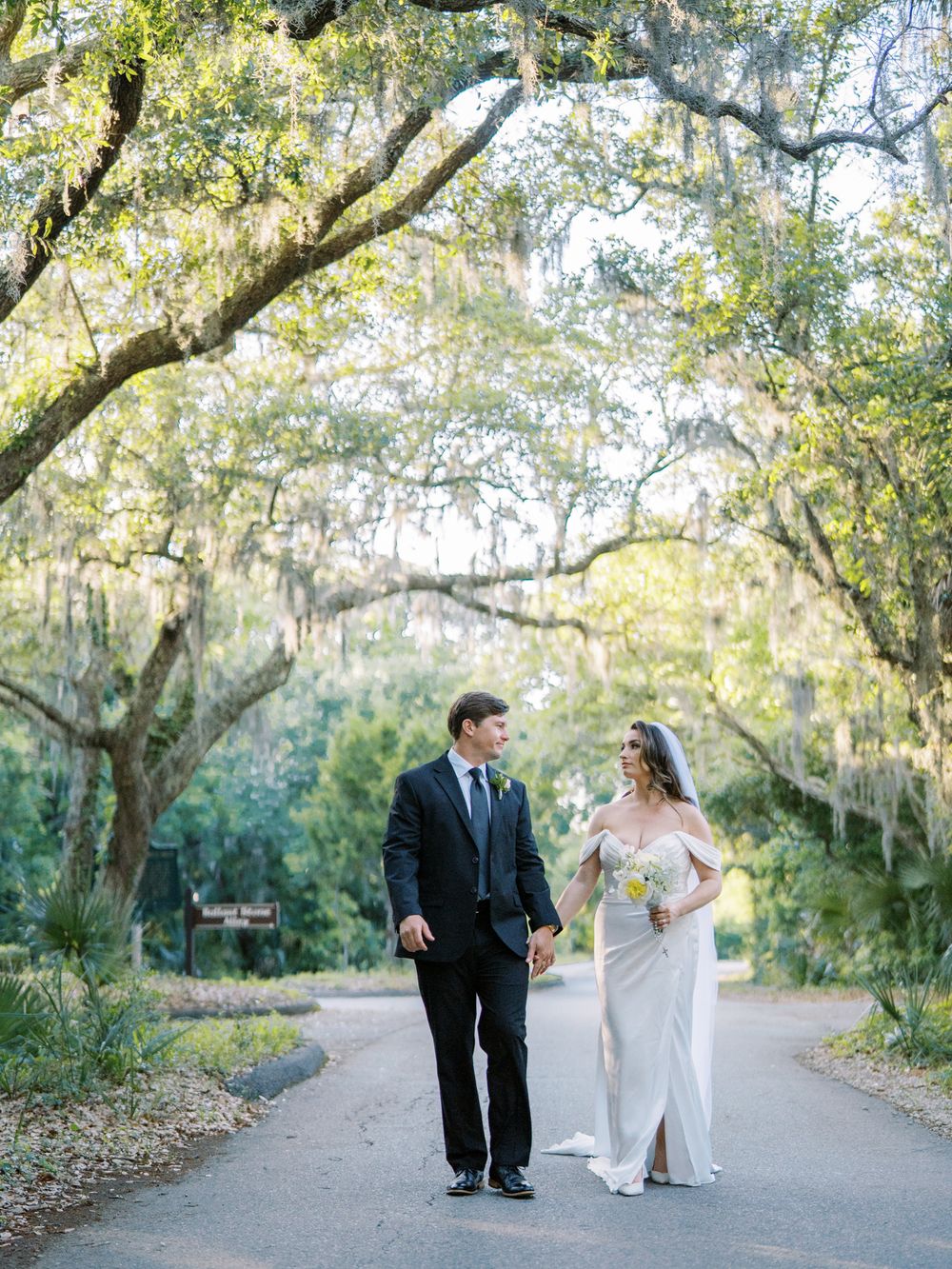 A couple walks down a tree-lined path with Spanish moss hanging from branches overhead on their wedding day.