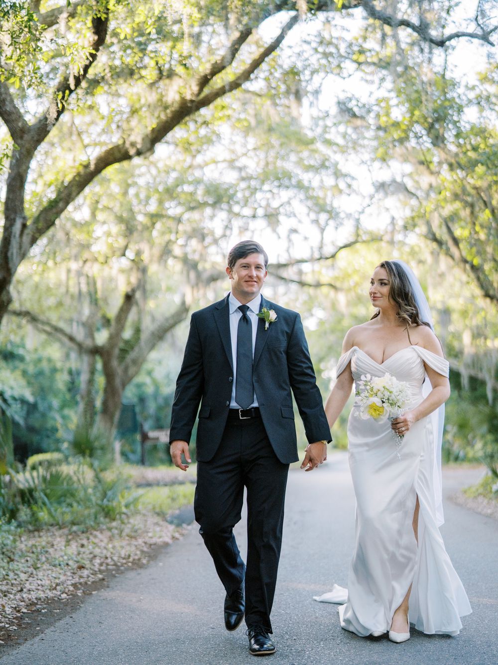 Wedding couple walking hand in hand on tree-lined path.