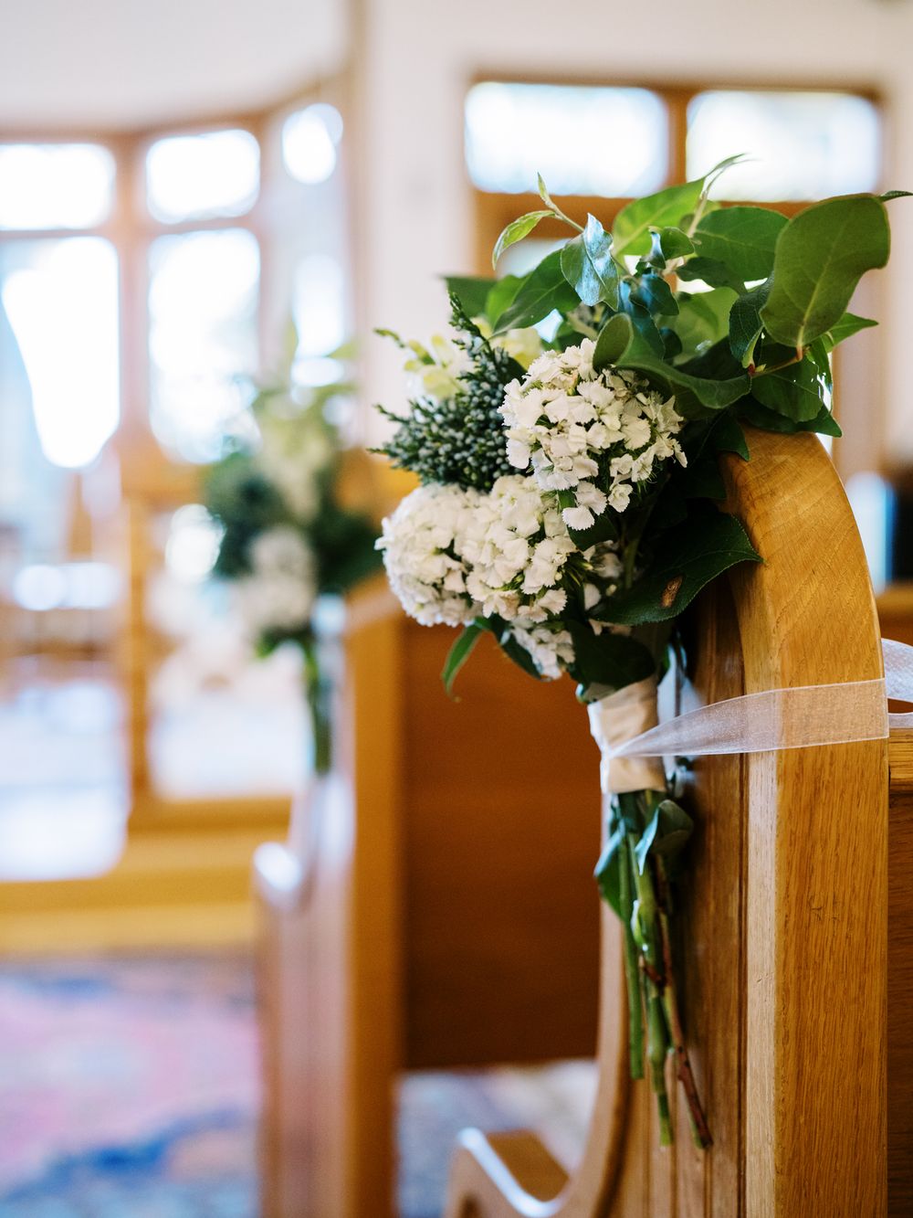 White floral pew decoration with greenery tied to wooden church pew.