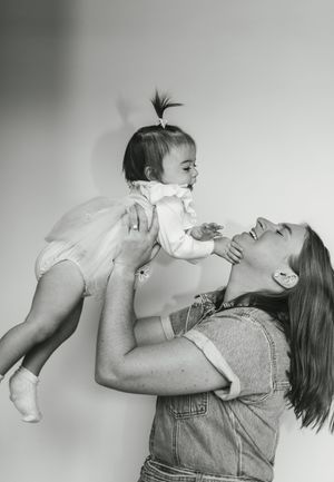 A parent in denim overalls lifts and plays with a baby wearing white, shown in a series of black and white photos.