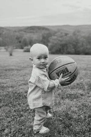 Black and white photo of a small child holding a basketball while standing in a grassy field with rolling hills in background.