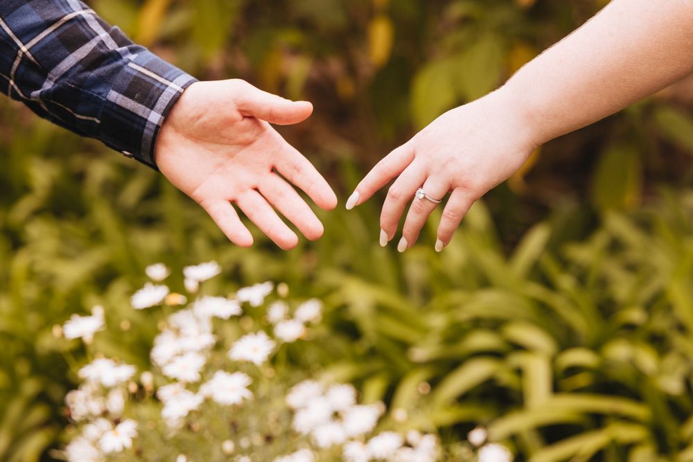 Close-up of hands with engagement ring against blurred floral background.