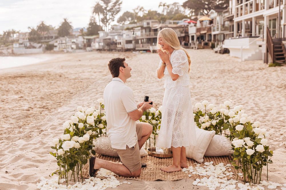 Man proposes to woman on beach with white roses and ring box on sandy shore near ocean.