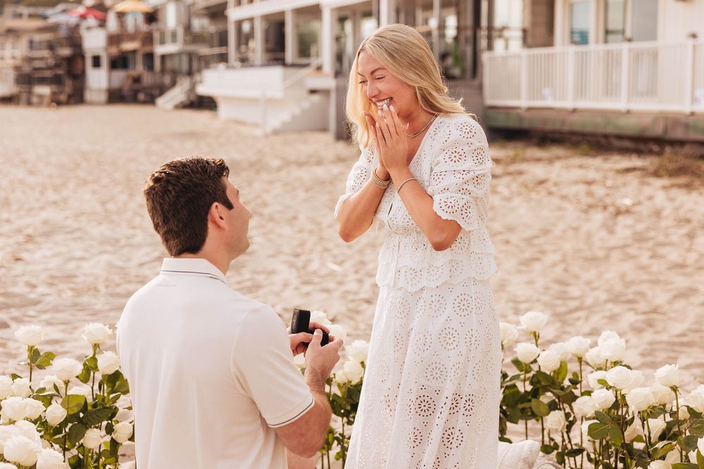 Man proposing on beach with ring box to surprised woman in white lace dress, surrounded by white roses.