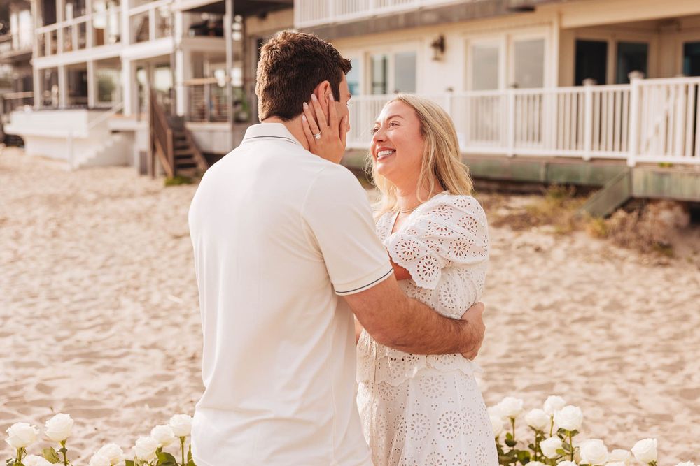 Couple in white outfits laughing together on a beach with white flowers, during a romantic proposal moment.