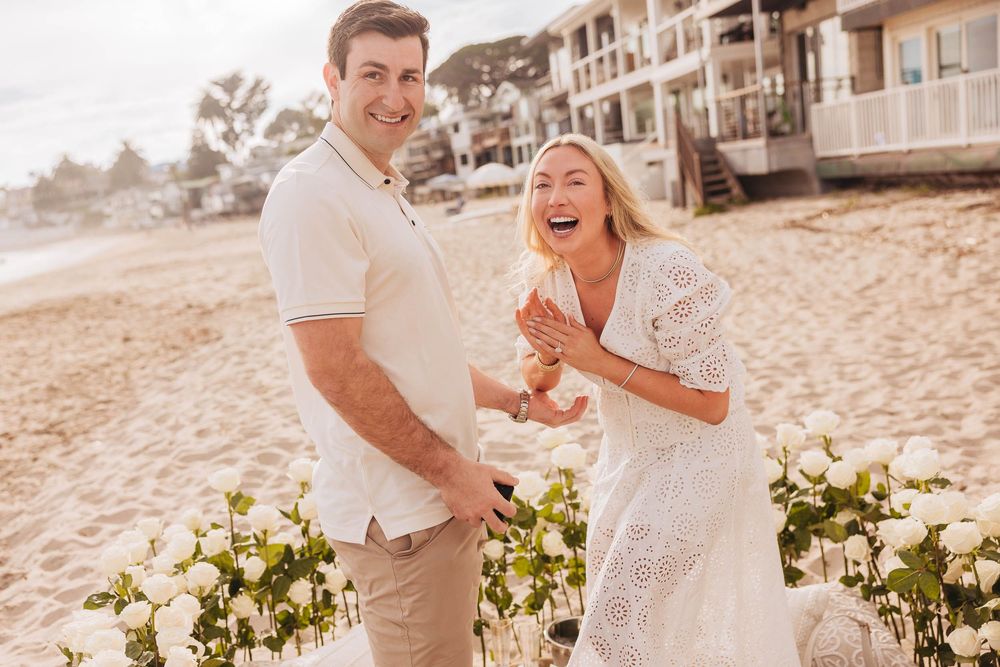 Happy couple laughing during beach proposal surrounded by white roses with ocean and resort in background.