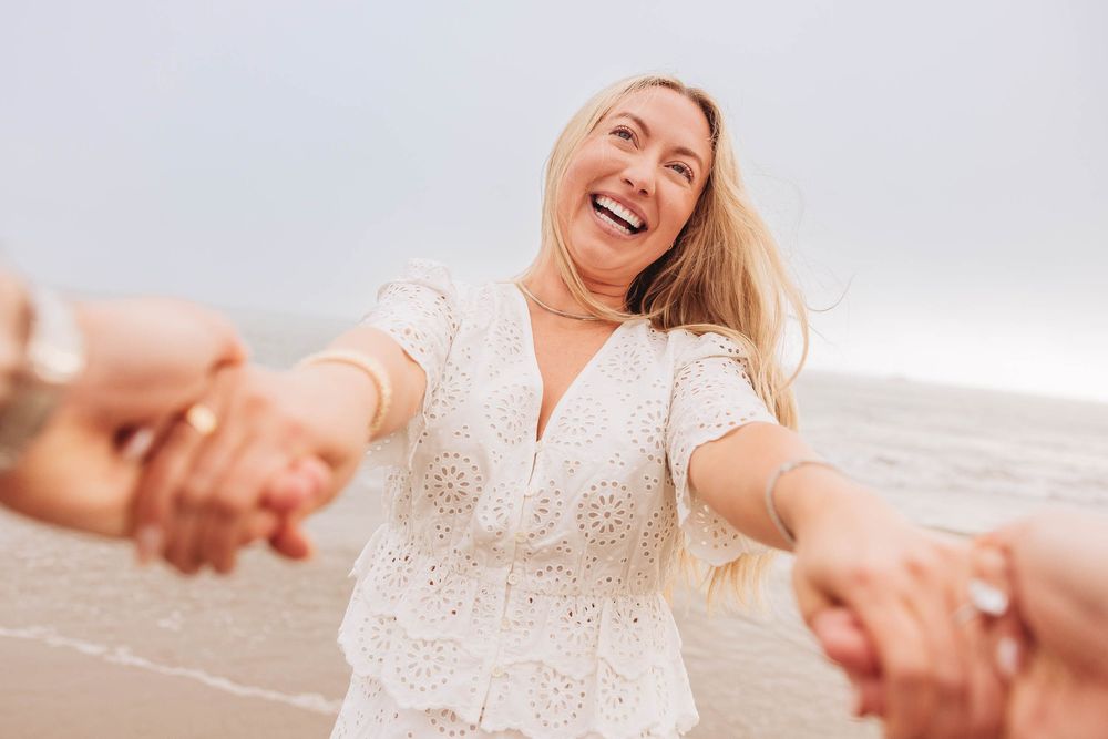A joyful blonde woman in a white lace dress laughs while holding hands with others on a beach.