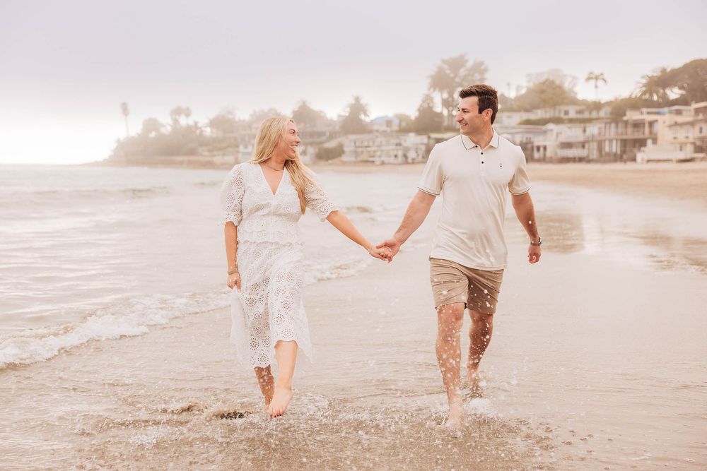 A couple walks hand-in-hand along a misty beach shore, smiling at each other in casual elegant attire.