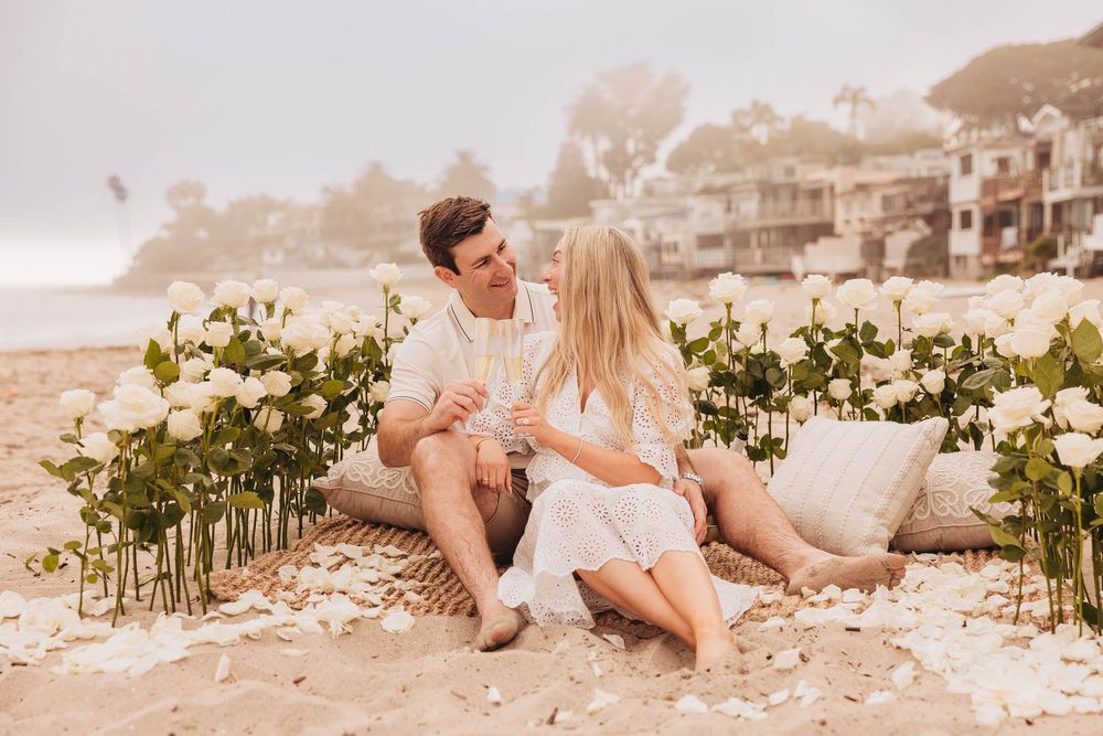 Couple sitting on a beach surrounded by white roses and flower petals during a romantic proposal setup.