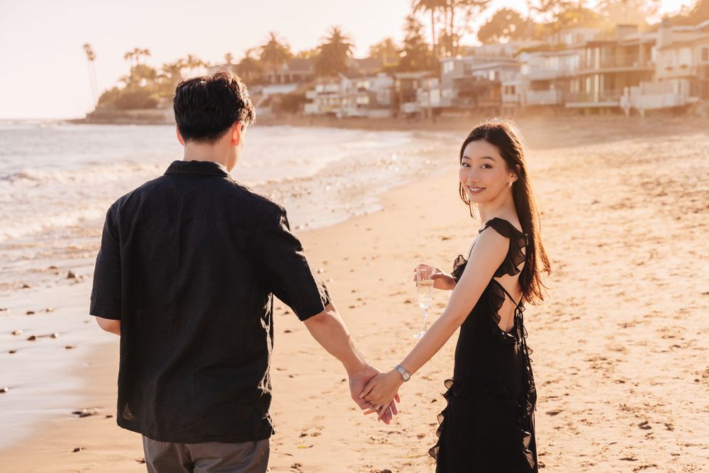 Couple holding hands while walking on beach at sunset with coastal homes in background.