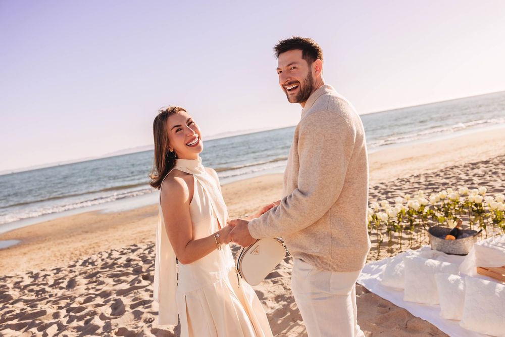 Happy couple holding hands and laughing on a sunny beach, dressed in elegant white and beige outfits.