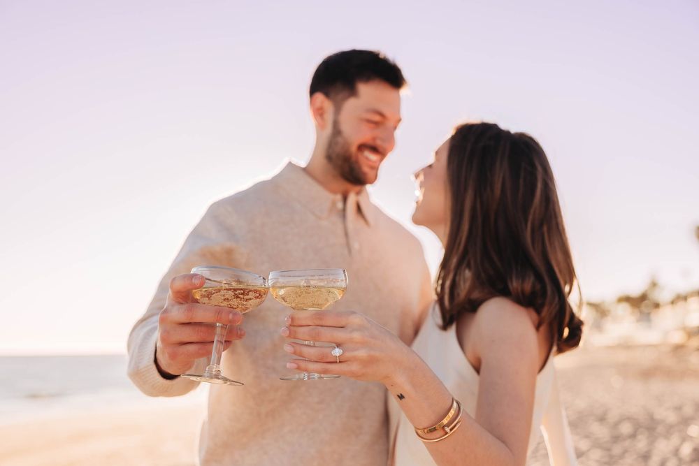 Couple toasting with champagne glasses on a beach at sunset, smiling at each other in elegant casual attire.