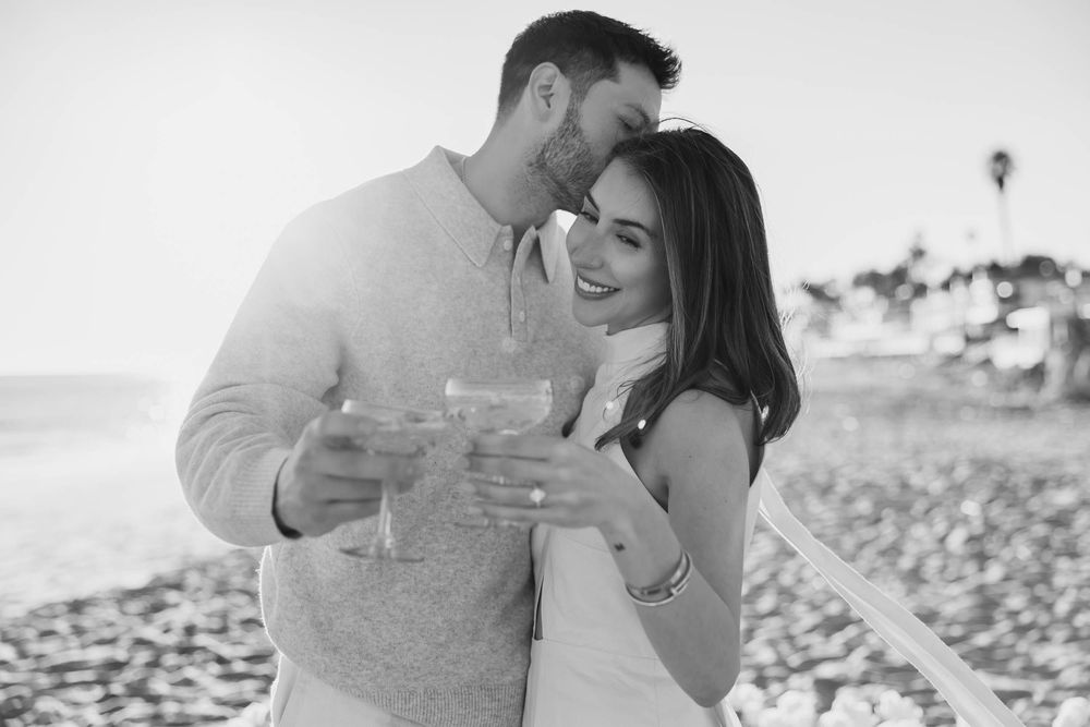 Black and white photo of a couple smiling and holding drinks together on a beach at sunset.