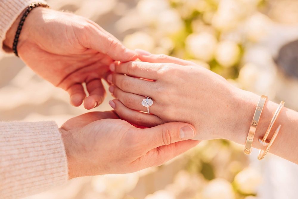 Close-up of a romantic engagement ring being placed on a woman's finger, with gold bracelets and soft floral bokeh background.