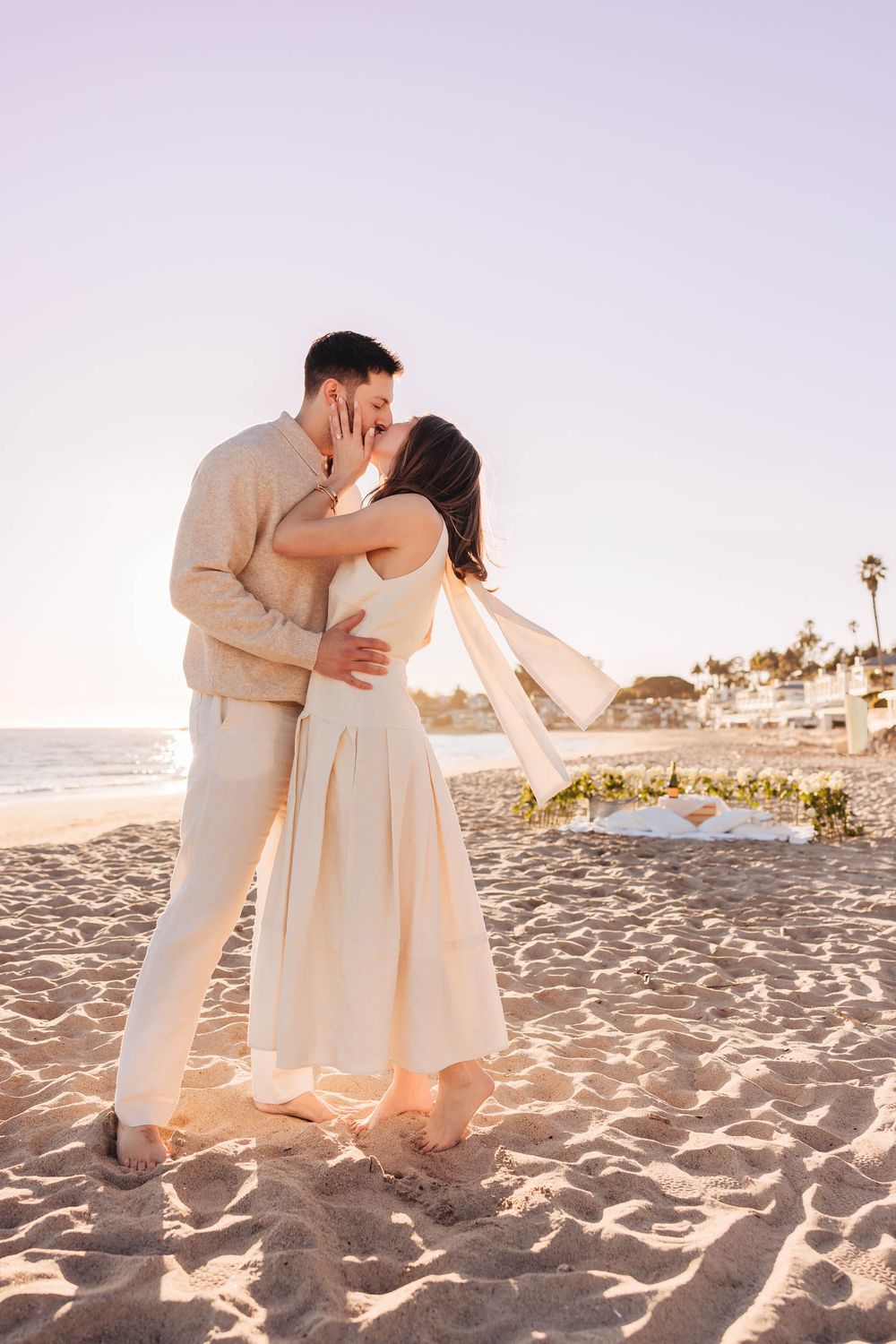 Couple sharing a romantic kiss on a sandy beach at sunset, dressed in matching cream outfits near a floral arrangement.
