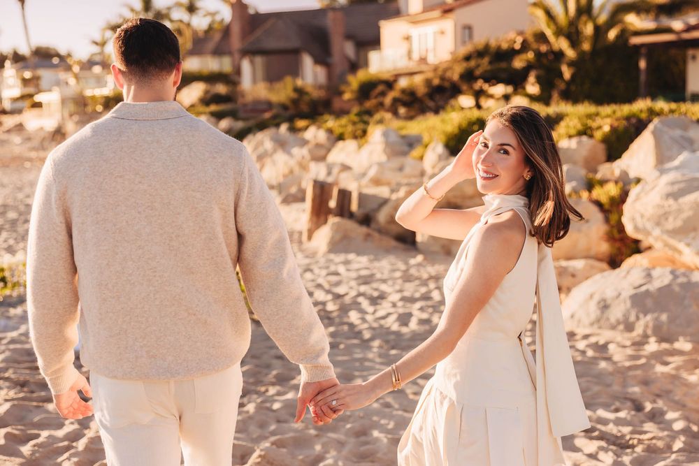 Couple holding hands on a sandy beach at sunset, woman in white dress smiling over her shoulder.