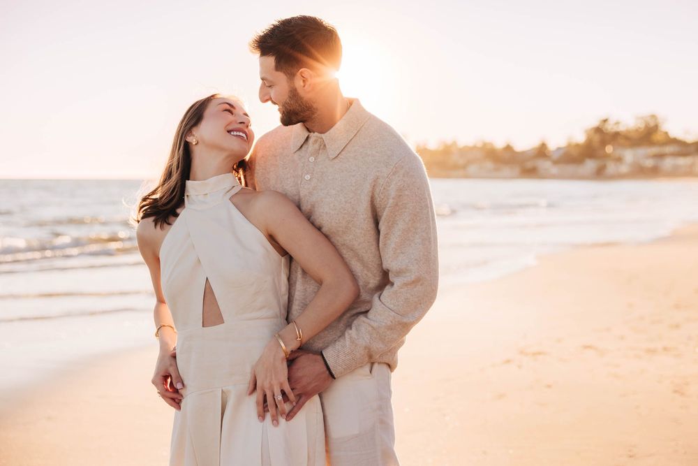 Couple embracing on a sunny beach at golden hour, woman smiling in white dress, man in beige shirt.