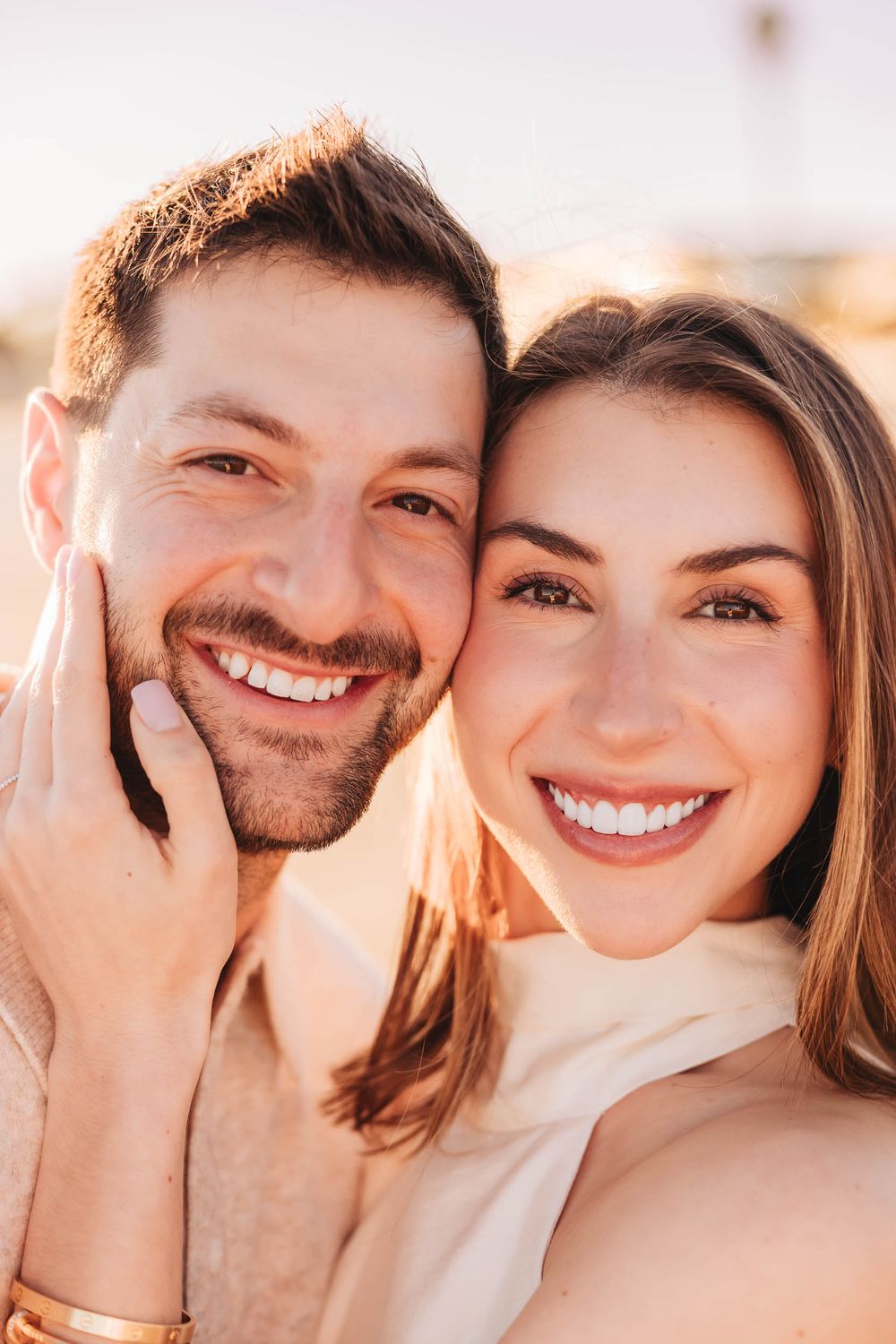 A smiling couple poses closely together in warm golden sunlight, the woman embracing the man from behind outdoors.