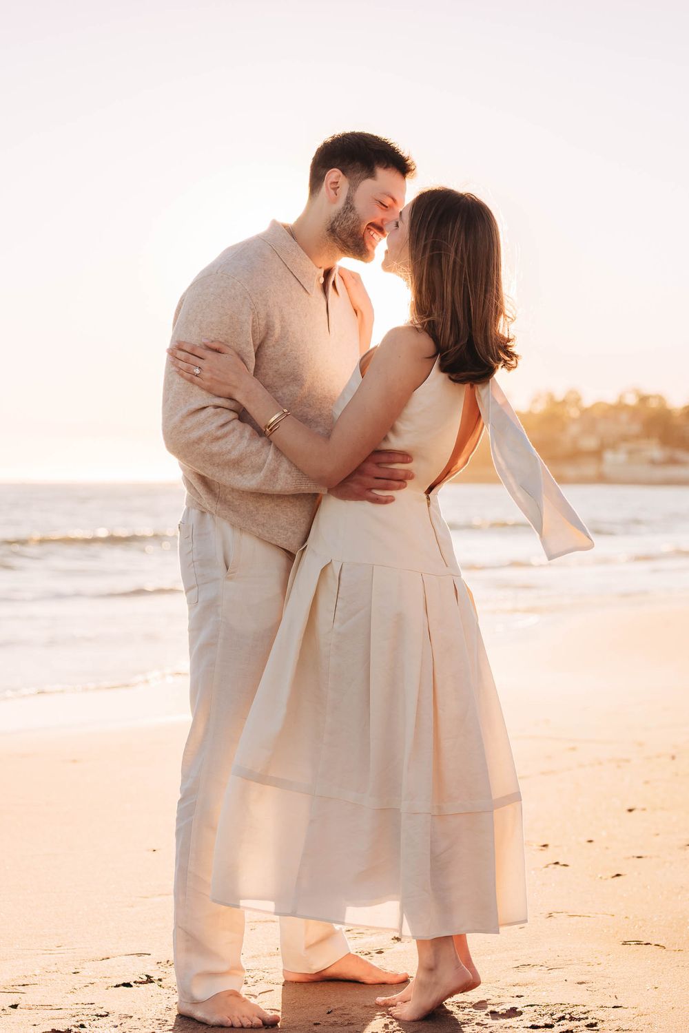 Couple embracing barefoot on a sandy beach at golden hour, wearing matching cream linen outfits with ocean waves behind them.