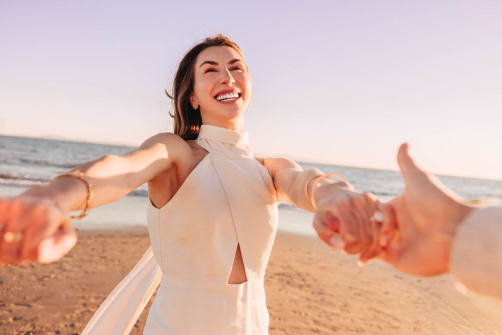 Joyful woman in white dress holding hands on sunny beach with ocean backdrop.