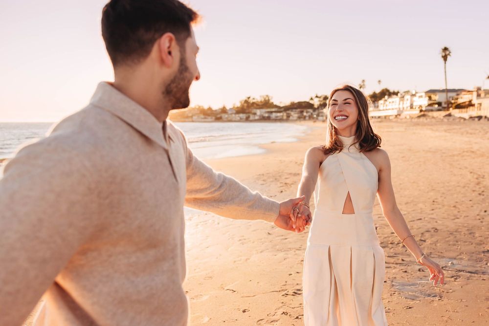 Couple holding hands on a sunny beach, woman in white dress smiling, golden hour light, romantic seaside moment.