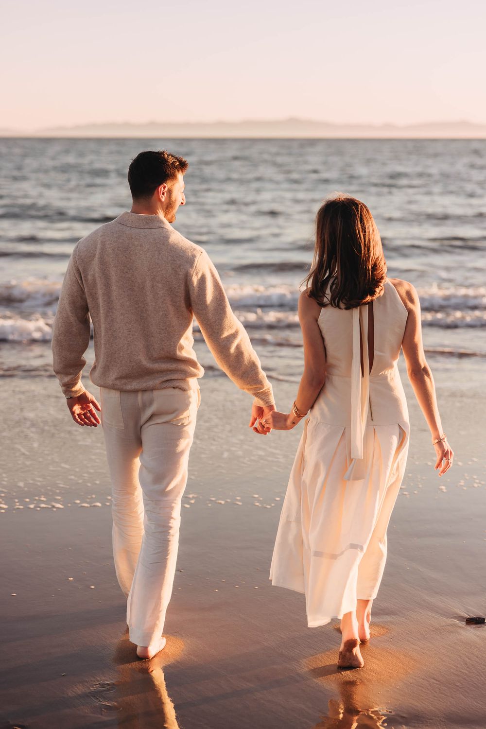 Couple holding hands walking along a sandy beach at sunset, wearing white and beige outfits.