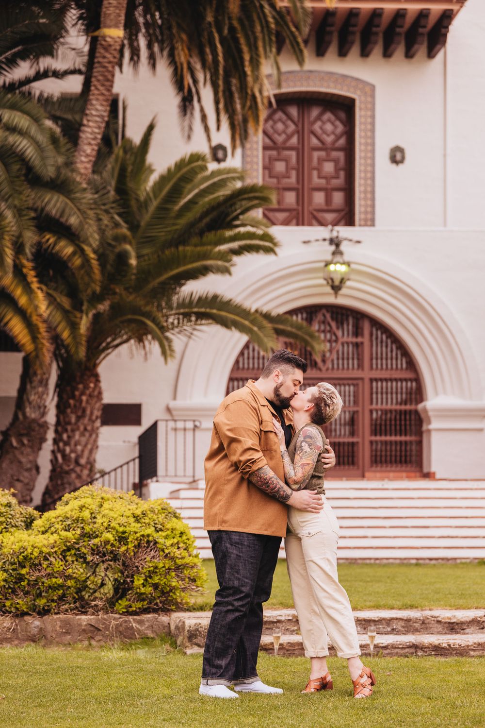 Couple celebrates engagement in front of Spanish colonial mission with palm trees.