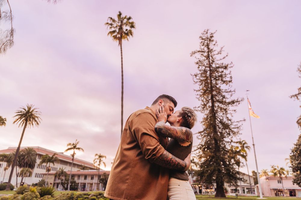 Romantic embrace under palm trees at sunset with Spanish architecture in background.