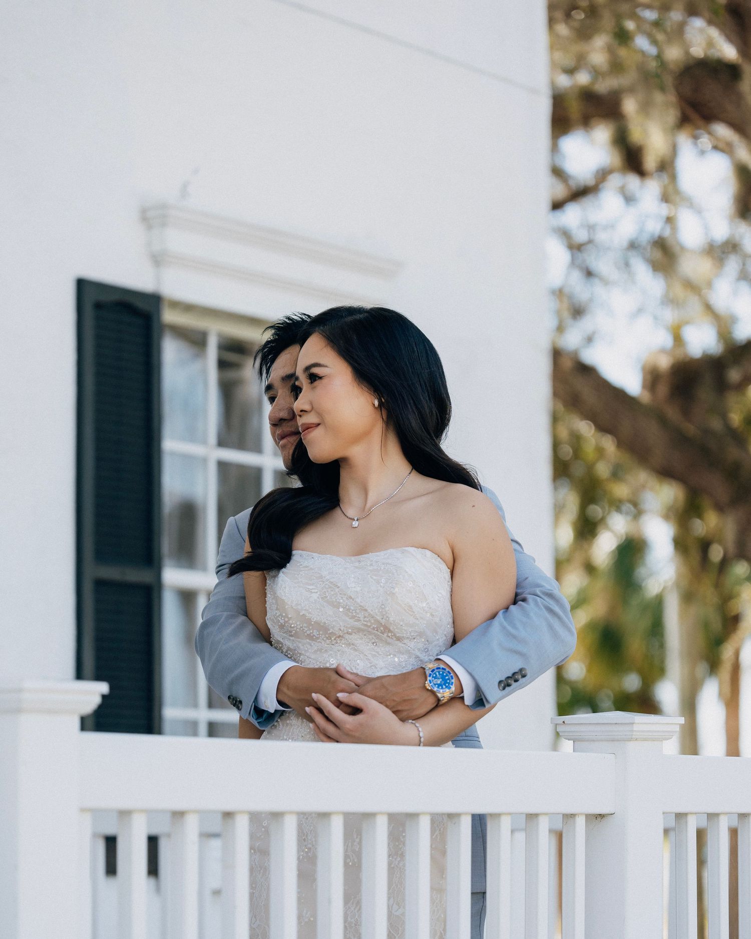 Bride in strapless lace gown and groom in gray suit embrace on a white porch balcony of a historic venue.