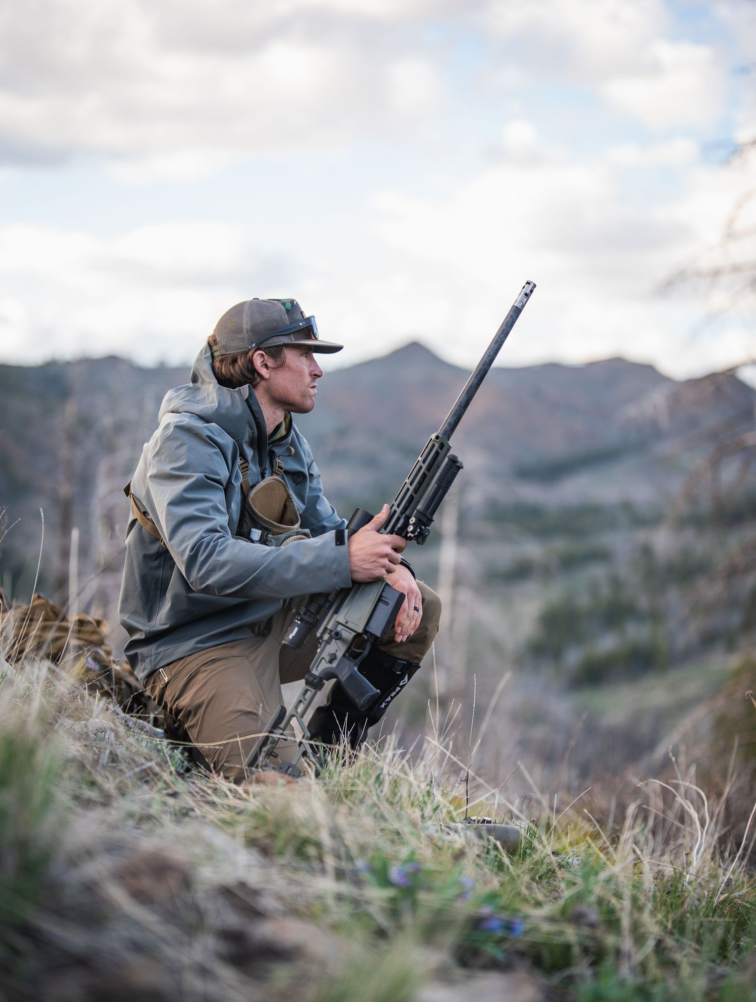 Hunter in tactical gear positioned on mountainside with rifle and equipment.