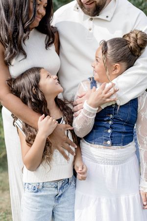 A joyful family of four in white outfits, two young girls laughing and hugging their parents outdoors.