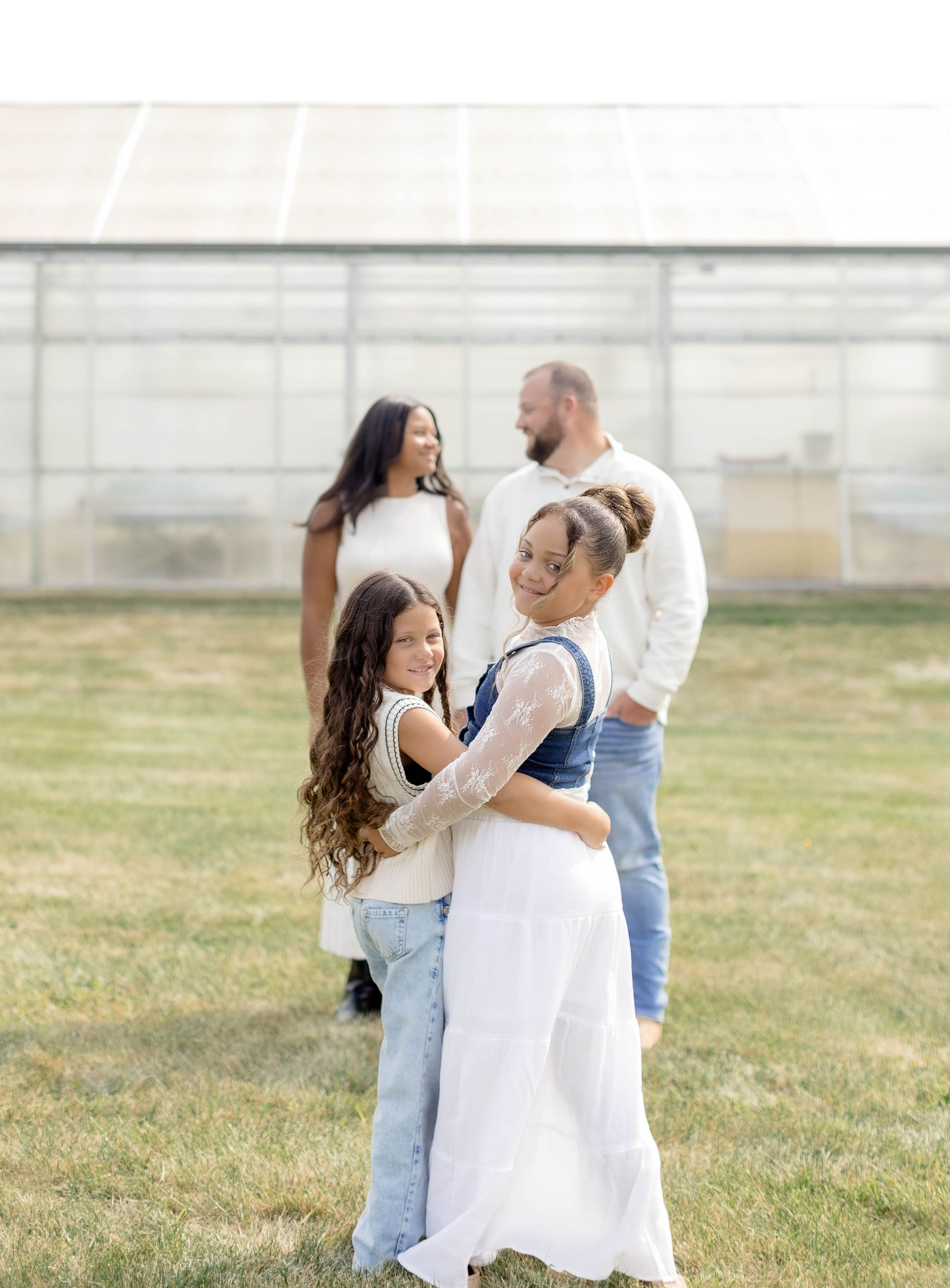 Family of four posing outdoors near a greenhouse, with two girls hugging in front while parents smile behind them.