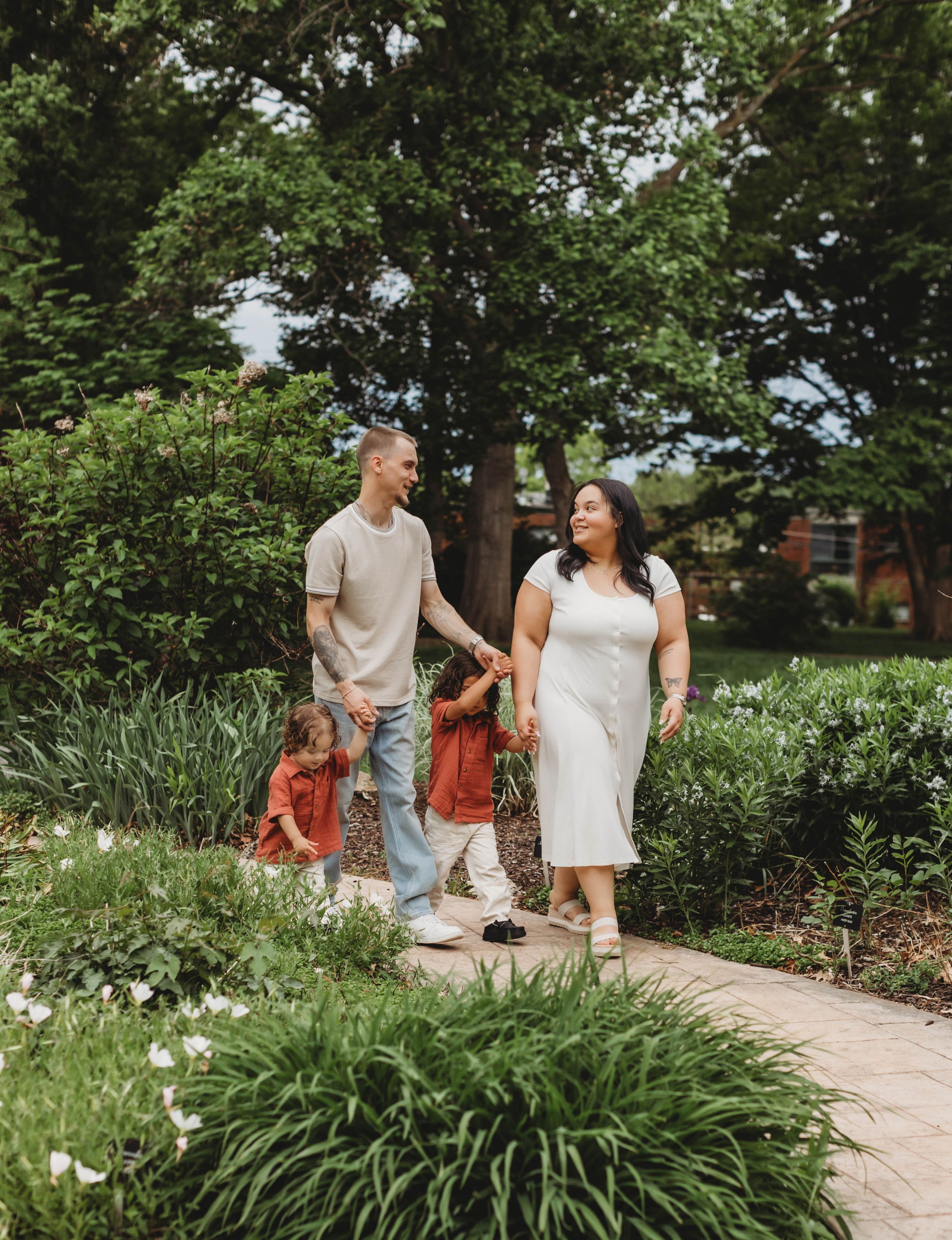 A happy family of four walks along a garden path surrounded by lush greenery, with parents holding hands with two young children.