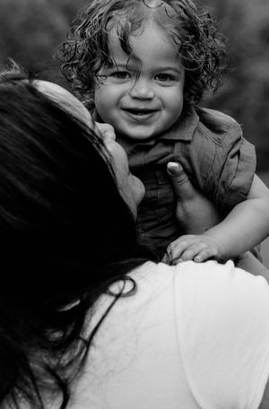 Black and white photo of a smiling curly-haired toddler being held up by a dark-haired woman in a white shirt.