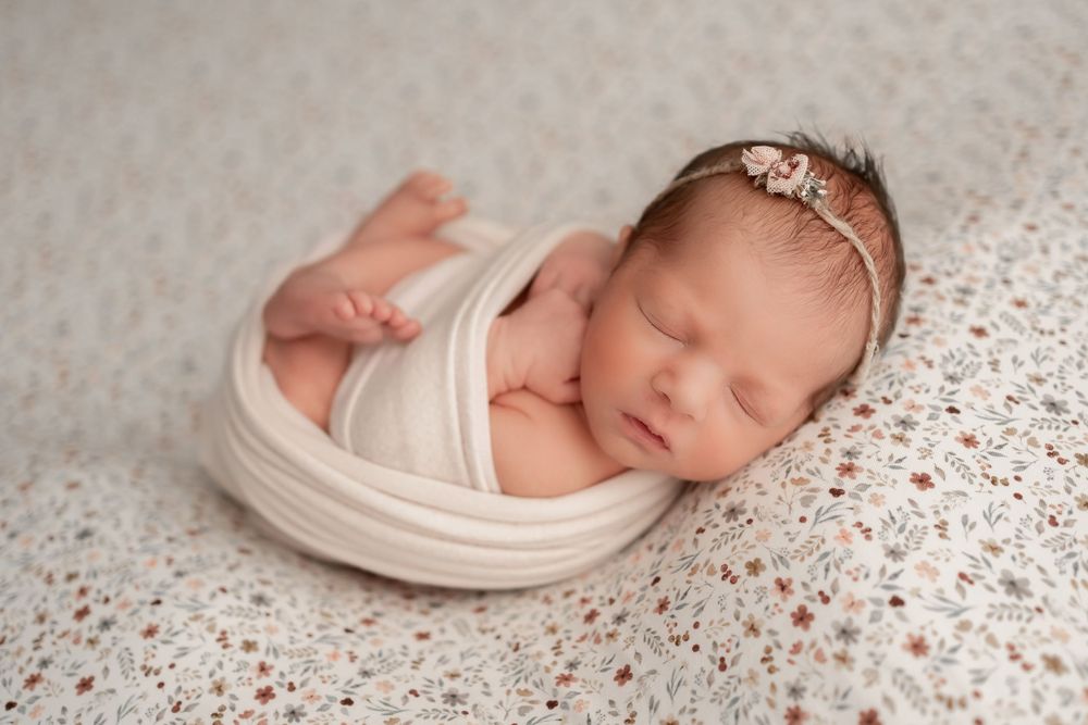 Sleeping newborn wrapped in white swaddle on speckled backdrop wearing delicate headband.