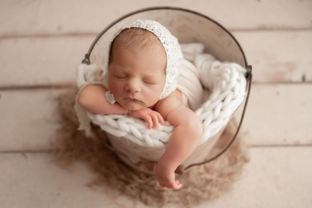 Sleeping newborn nestled in white knit blanket inside rustic metal bowl.